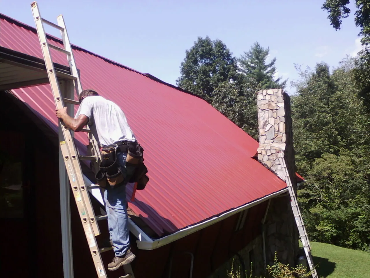 Roofer on a ladder beside a red metal roof and stone chimney — Soffit & Fascia Repair by Roofing Brothers in Codorus